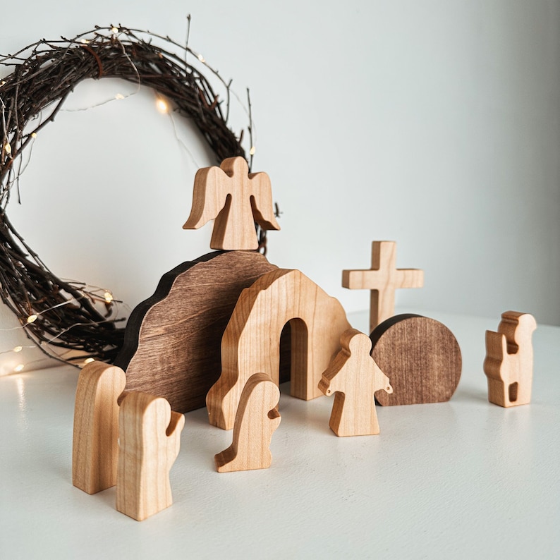 Wooden Easter resurrection scene with Jesus, angel, empty tomb, and cross, handcrafted from natural wood. Minimalist Christian decor with rustic wreath and warm lights, creating a cozy faith-inspired display. Montessori-style wooden toy set with smooth finish, perfect for Easter decor, baptism gift, baby shower, or meaningful religious keepsake for kids.
