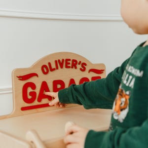 May include: A light-colored wooden sign with a curved top edge featuring the red text "OLIVER'S GARAGE" in a bold, sans-serif font. The sign includes decorative red swoosh accents on either side of the word Oliver's and a horizontal line.