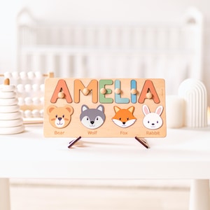 Puede incluir: Rompecabezas de madera con el nombre "AMELIA" en letras coloridas. El rompecabezas presenta ilustraciones de un oso, un lobo, un zorro y un conejo. El rompecabezas est&aacute; dise&ntilde;ado para ni&ntilde;os y se muestra sobre una superficie blanca.