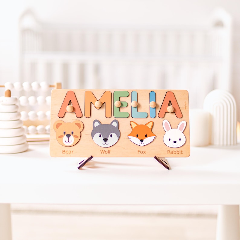 Puede incluir: Rompecabezas de madera con el nombre "AMELIA" en letras coloridas. El rompecabezas presenta ilustraciones de un oso, un lobo, un zorro y un conejo. El rompecabezas est&aacute; dise&ntilde;ado para ni&ntilde;os y se muestra sobre una superficie blanca.