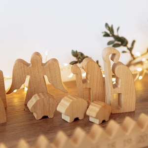Pu&ograve; includere: Un set di statuine per presepe in legno chiaro che include un angelo con le ali spiegate, un pastore con un bastone e tre piccole pecore. Le figure in legno liscio e intagliato sono disposte in gruppo su una superficie piana.