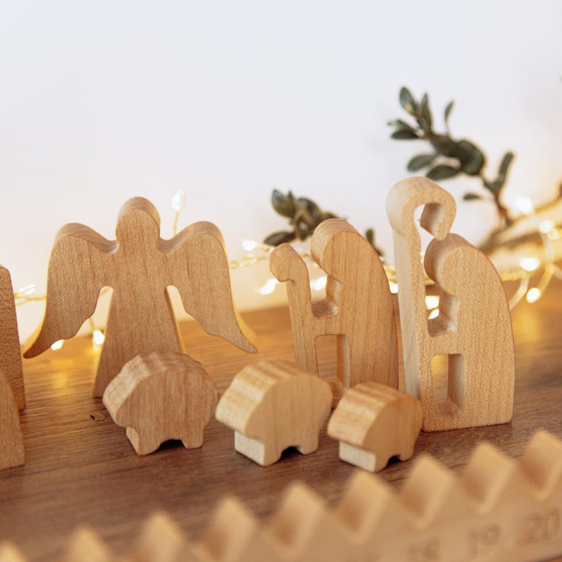 Pu&ograve; includere: Un set di statuine per presepe in legno chiaro che include un angelo con le ali spiegate, un pastore con un bastone e tre piccole pecore. Le figure in legno liscio e intagliato sono disposte in gruppo su una superficie piana.