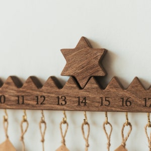 Pu&ograve; includere: Un calendario da parete in legno marrone scuro con un bordo superiore seghettato e un ornamento a forma di stella a sei punte. La barra orizzontale in legno &egrave; incisa con i numeri da 10 a 17 e include piccoli ganci in ottone con lacci.