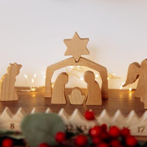 Pu&ograve; includere: Un presepe in legno con figure stilizzate di due persone e un bambino sotto un arco a forma di stalla sormontato da una stella. Ai lati si trovano ulteriori figure in legno, tra cui un angelo, e una base in legno numerata.