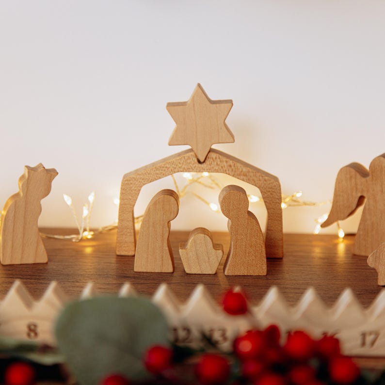 Pu&ograve; includere: Un presepe in legno con figure stilizzate di due persone e un bambino sotto un arco a forma di stalla sormontato da una stella. Ai lati si trovano ulteriori figure in legno, tra cui un angelo, e una base in legno numerata.