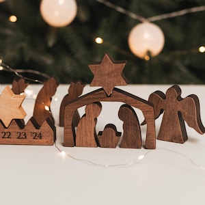 Pu&ograve; includere: Un presepe in legno marrone scuro composto da una struttura centrale a forma di stalla sormontata da una stella, figure di due adulti e un bambino, e un angelo separato. A sinistra, una base in legno mostra i numeri 22, 23 e 24.