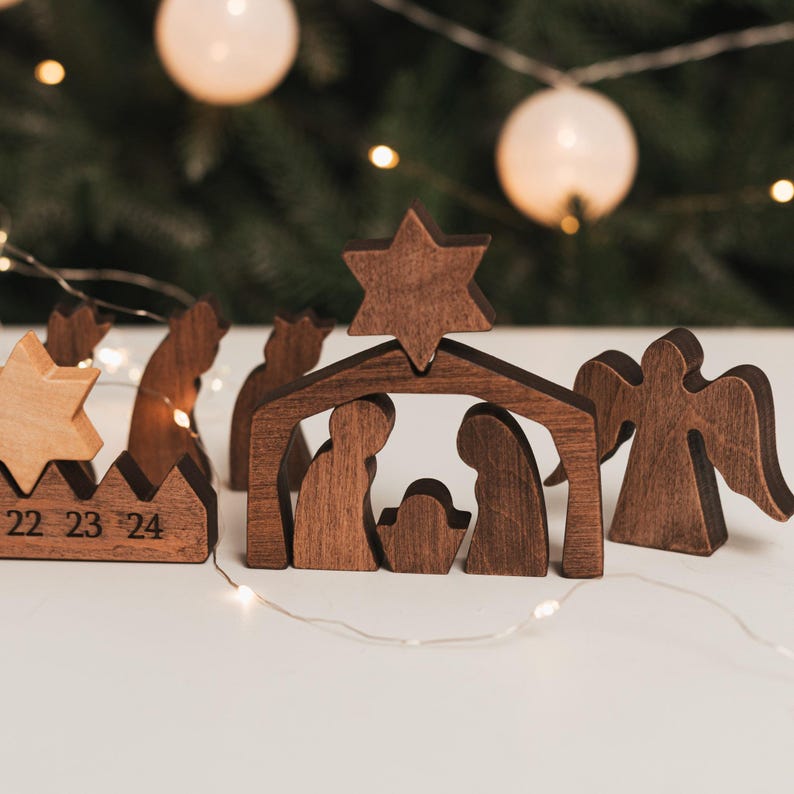 Pu&ograve; includere: Un presepe in legno marrone scuro composto da una struttura centrale a forma di stalla sormontata da una stella, figure di due adulti e un bambino, e un angelo separato. A sinistra, una base in legno mostra i numeri 22, 23 e 24.