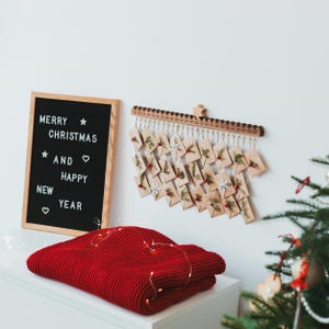 Pu&ograve; includere: Una lavagna in feltro nero con cornice in legno che mostra il testo MERRY CHRISTMAS AND HAPPY NEW YEAR, insieme a un calendario dell'avvento da parete in legno con venticinque piccole buste decorate con vegetazione e bacche rosse.