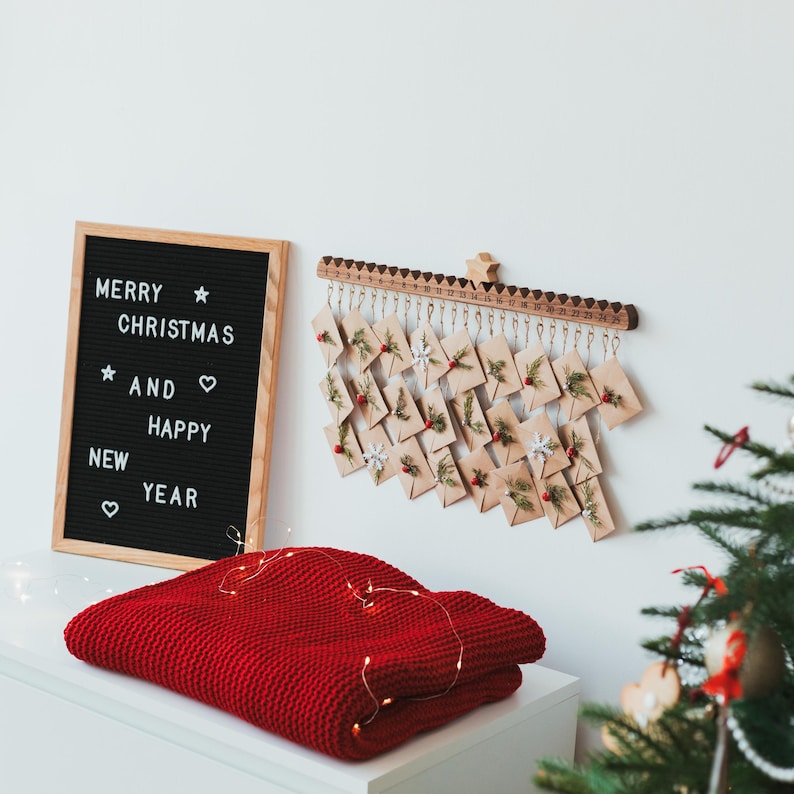 Pu&ograve; includere: Una lavagna in feltro nero con cornice in legno che mostra il testo MERRY CHRISTMAS AND HAPPY NEW YEAR, insieme a un calendario dell'avvento da parete in legno con venticinque piccole buste decorate con vegetazione e bacche rosse.
