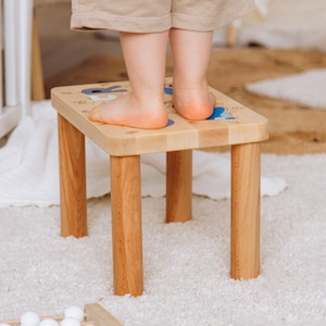 May include: A light-colored wooden step stool with four cylindrical legs and a rectangular top surface. The top of the stool features blue and white printed text and small graphic illustrations, providing a stable platform for standing.