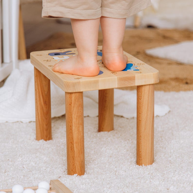 May include: A light-colored wooden step stool with four cylindrical legs and a rectangular top surface. The top of the stool features blue and white printed text and small graphic illustrations, providing a stable platform for standing.