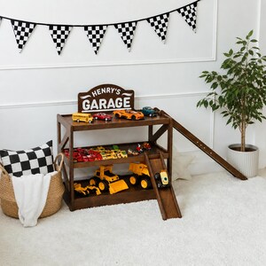 May include: A dark brown wooden toy garage with three levels, a ramp, and a sign reading "HENRY'S GARAGE". The shelves are filled with various small toy cars and yellow construction vehicles. A checkered flag banner hangs above the unit.