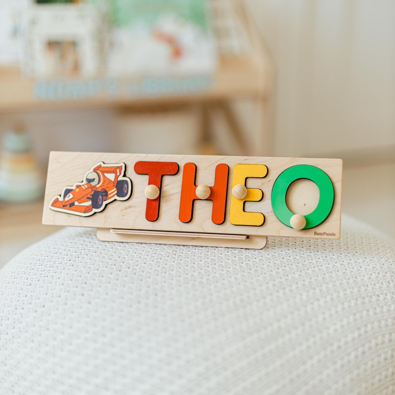 以下が含まれることがあります： 赤のT、オレンジのH、黄色のE、緑のOというカラフルな文字で「THEO」と書かれた木製の名前パズル。各文字には掴むための小さな木製のノブが付いています。名前の左側にはオレンジ色のレーシングカーのイラストが描かれています。