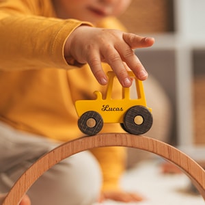 Puede incluir: Un tractor de juguete de madera amarillo con ruedas de color marr&oacute;n oscuro y el nombre Lucas impreso en letra cursiva negra en el lateral, colocado sobre una pieza de arco de madera curvada. El tractor tiene un dise&ntilde;o simple con una chimenea.