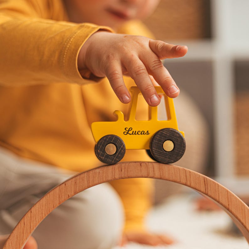 Puede incluir: Un tractor de juguete de madera amarillo con ruedas de color marr&oacute;n oscuro y el nombre Lucas impreso en letra cursiva negra en el lateral, colocado sobre una pieza de arco de madera curvada. El tractor tiene un dise&ntilde;o simple con una chimenea.