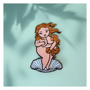 May include: A pin featuring a cartoon illustration of a nude woman with red hair, standing on a seashell. The woman is depicted with a curvy figure and is partially covering her breasts. The pin has a gold outline and is set against a light blue background.