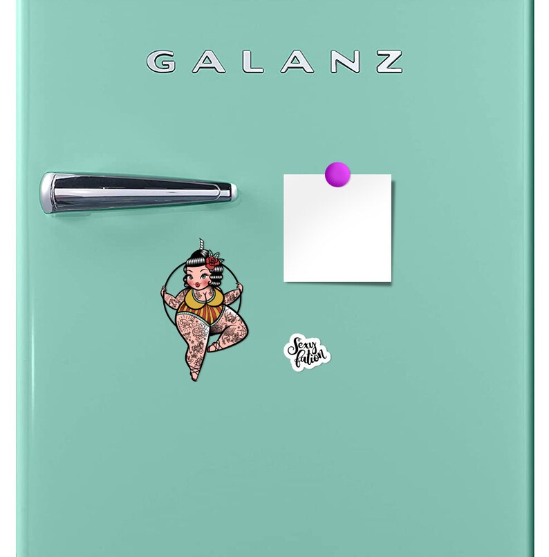 May include: A mint green refrigerator with the brand name "GALANZ" in silver lettering. A chrome handle and a purple magnet hold a blank white square. A sticker with a pin-up girl and the text "Sexy Fation" is also on the refrigerator.