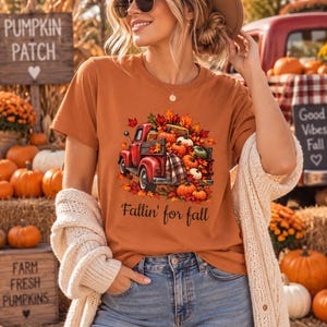 May include: A burnt orange short-sleeve t-shirt featuring a graphic print of a red pickup truck filled with pumpkins and autumn leaves, with the text Fallin' for fall printed in black script lettering below the truck illustration.