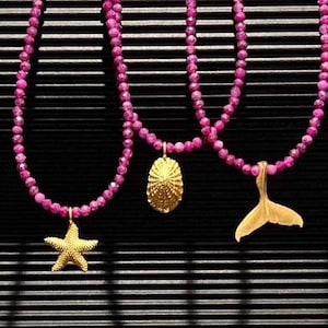 Può includere: Tre collane con perline di pietre preziose rosa e ciondoli in oro. I ciondoli sono una stella marina, una conchiglia e una coda di balena.