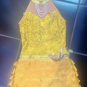May include: A yellow sleeveless dress featuring a sequined bodice, a high collar with a faux pearl necklace detail, and a skirt layered with multiple rows of fringe. A decorative gold sequin appliqué with yellow feathers sits on the hip.
