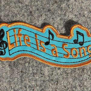 May include: Embroidered patch with the text "Life is a Song" in orange, set against a light blue background with musical notes and staff lines. The patch has an orange border and a black treble clef.