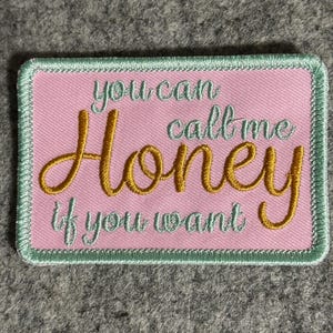 Könnte beinhalten: Ein rechteckiger rosa Aufnäher mit den Worten "you can call me Honey if you want" in Gold und Hellgrün gestickt. Der Aufnäher hat einen hellgrünen Rand.