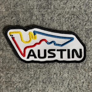 May include: Embroidered patch featuring a stylized racetrack design with red, yellow, and blue lines on a white background. The word "AUSTIN" is embroidered in black capital letters. The patch has a black border.