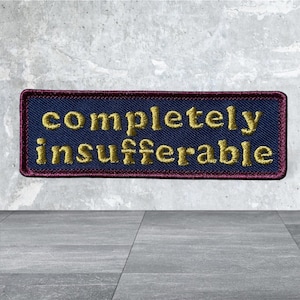 May include: A rectangular embroidered patch featuring a dark blue background with a deep red border. The text "completely insufferable" is stitched in gold thread across the center in a clear, sans-serif font style.