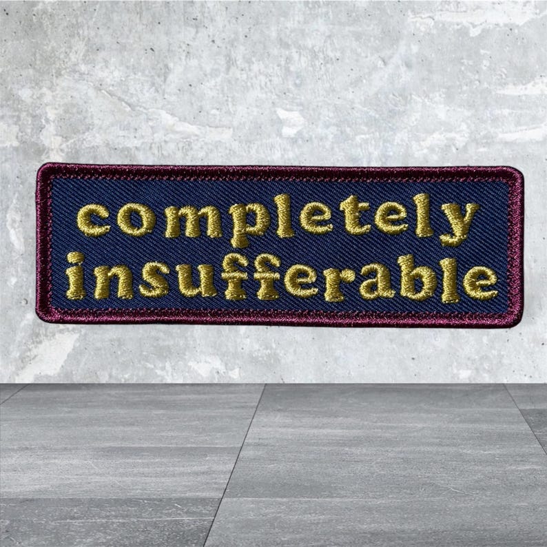 Peut inclure: Un &eacute;cusson rectangulaire brod&eacute; avec un fond bleu fonc&eacute; et une bordure rouge fonc&eacute;. Le texte "completely insufferable" est cousu en fil dor&eacute; au centre dans une police de caract&egrave;res claire et sans empattement.