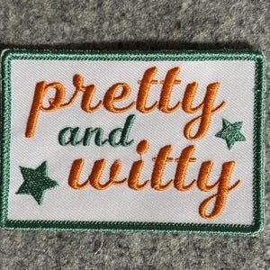 Könnte beinhalten: Gestickter Aufnäher mit den Worten "pretty and witty" in orangefarbener Schrift auf weißem Hintergrund. Der Aufnäher hat einen grünen Rand und grüne Sterne.