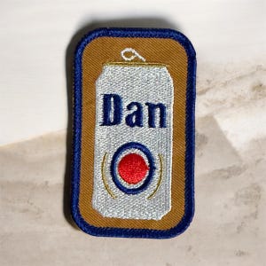 May include: Embroidered patch featuring a white beer can design with the name "Dan" in blue lettering. The can has a red and blue circular emblem. The patch is bordered by a blue and brown rectangle.