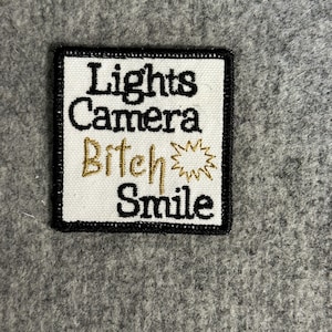 May include: White embroidered patch with black border. The text reads "Lights Camera Bitch Smile" with a gold starburst.