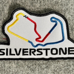 May include: A white embroidered patch with a black border featuring a stylized map of the Silverstone race circuit in yellow, red, and blue thread. The word SILVERSTONE is stitched in bold black capital letters at the bottom of the patch.