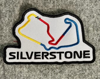 Peut inclure: Un écusson brodé blanc avec une bordure noire représentant une carte stylisée du circuit de Silverstone en fil jaune, rouge et bleu. Le mot SILVERSTONE est brodé en lettres capitales noires et grasses au bas de l'écusson.