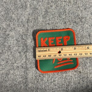 May include: A square patch with the word "KEEP" in orange letters on a green background, outlined in orange. The patch is approximately 3 inches wide.