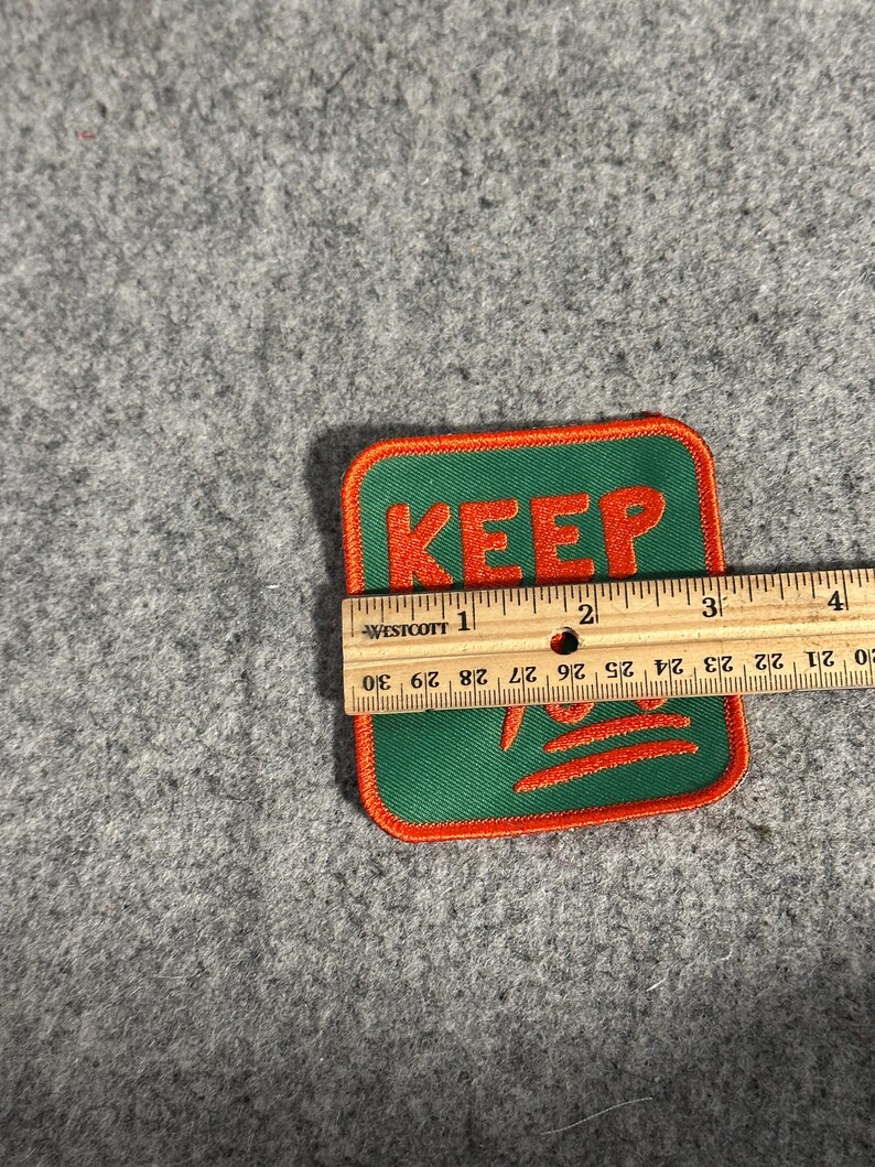 May include: A square patch with the word "KEEP" in orange letters on a green background, outlined in orange. The patch is approximately 3 inches wide.