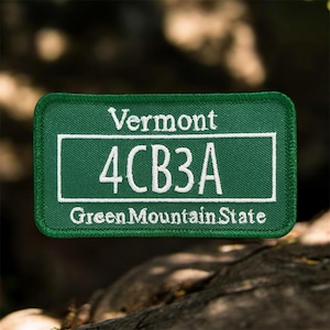 May include: A green rectangular patch with white embroidered text. The patch reads "Vermont" at the top, "4CB3A" in the center, and "Green Mountain State" at the bottom. The patch has a white border and a green stitched edge.