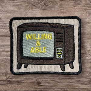May include: A rectangular embroidered patch featuring a vintage brown television set on a light beige background. The screen displays the yellow text WILLING & ABLE. The patch has a thick black border and four small legs at the bottom corners.
