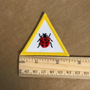 May include: Embroidered patch in a white triangle with a yellow border. The patch features a red ladybug with black spots and legs. The patch is approximately 5 cm wide.