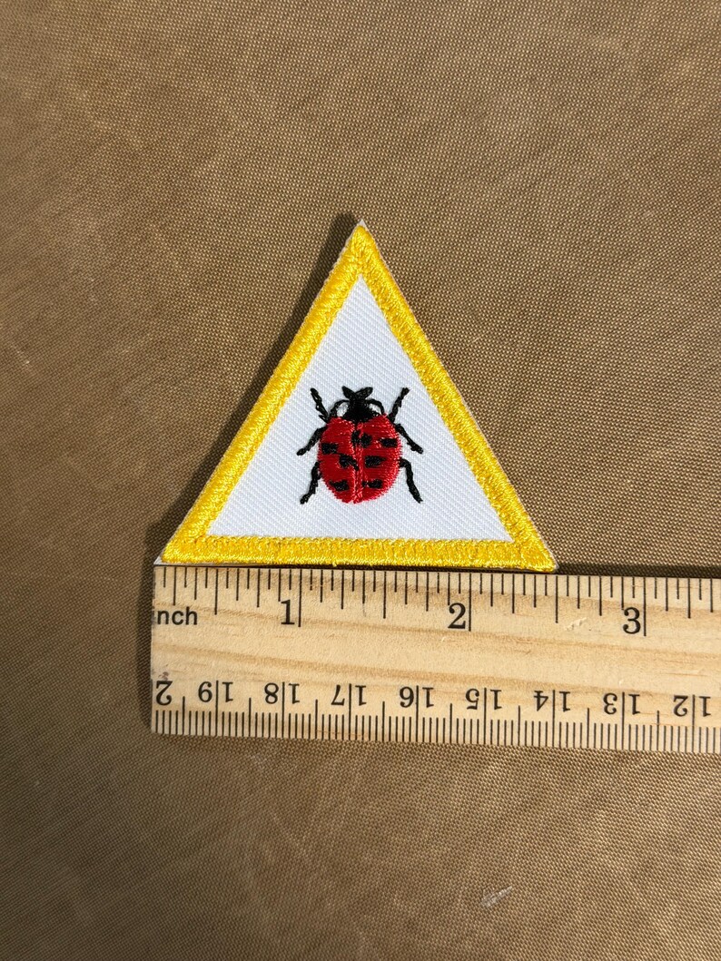 May include: Embroidered patch in a white triangle with a yellow border. The patch features a red ladybug with black spots and legs. The patch is approximately 5 cm wide.