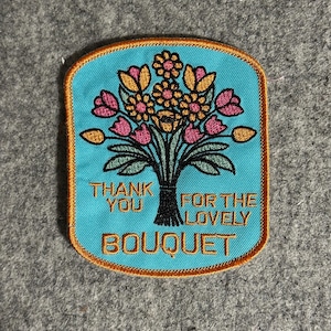 May include: Embroidered patch with a turquoise background and a bouquet of flowers in pink, yellow, and green. The patch has a gold border and the text "THANK YOU FOR THE LOVELY BOUQUET".