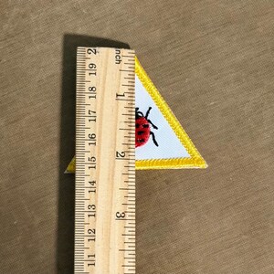 May include: A triangular patch with a white background and a yellow border. The patch features a red ladybug with black spots. The patch is approximately 5 cm in height, as measured by a wooden ruler.