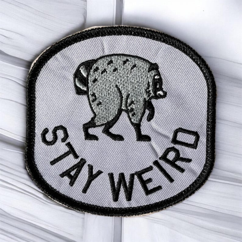 May include: A circular embroidered patch with a light gray background and a thick black border. The patch features a dark gray raccoon illustration and the black embroidered text STAY WEIRD curved along the bottom edge of the circular design.