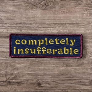 Peut inclure: Un &eacute;cusson rectangulaire brod&eacute; avec un fond en tissu bleu fonc&eacute; et une bordure rouge fonc&eacute;. Le texte "completely insufferable" est cousu en fil dor&eacute; au centre dans une police sans empattement en minuscules et en gras.