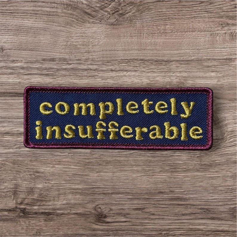 Peut inclure: Un &eacute;cusson rectangulaire brod&eacute; avec un fond en tissu bleu fonc&eacute; et une bordure rouge fonc&eacute;. Le texte "completely insufferable" est cousu en fil dor&eacute; au centre dans une police sans empattement en minuscules et en gras.