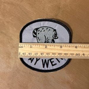 May include: A light gray embroidered patch with a black border featuring a graphic of a bear and the text "AY WE". A wooden ruler is placed across the patch, showing a width of approximately 3.5 inches for the embroidered design.