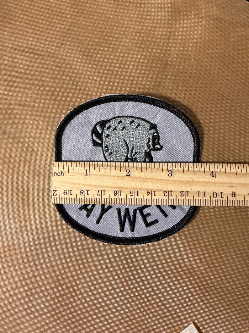 May include: A light gray embroidered patch with a black border featuring a graphic of a bear and the text "AY WE". A wooden ruler is placed across the patch, showing a width of approximately 3.5 inches for the embroidered design.