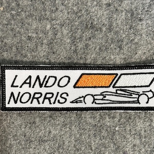 May include: A rectangular patch with the words "LANDO NORRIS" in black, above a line drawing of a race car. The patch has a white background with a black border and orange and white rectangles.