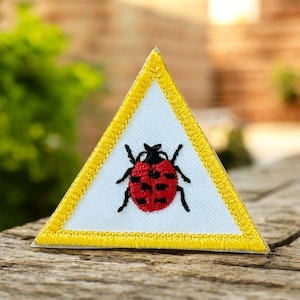 May include: A triangular patch featuring a white background, a yellow border, and a red ladybug design. The ladybug has black spots and legs. This embroidered patch could be for clothing or accessories.