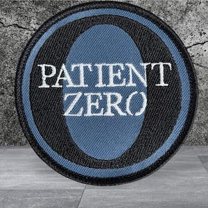 May include: A circular embroidered patch featuring a black border and a blue oval center. The text PATIENT ZERO is stitched in white capital letters across the middle of the blue oval. The design is flat with a visible fabric texture throughout.
